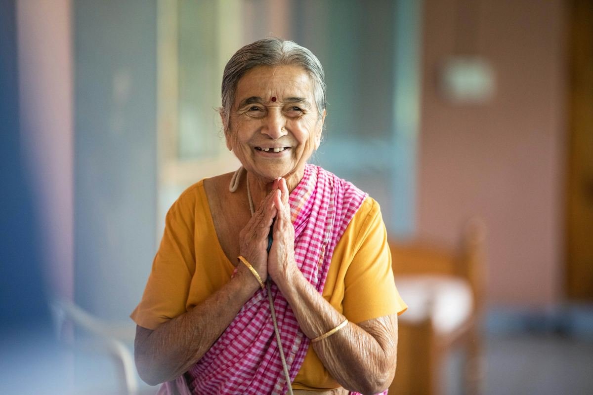 An older woman in a sari smiling warmly with her hands joined in greeting.